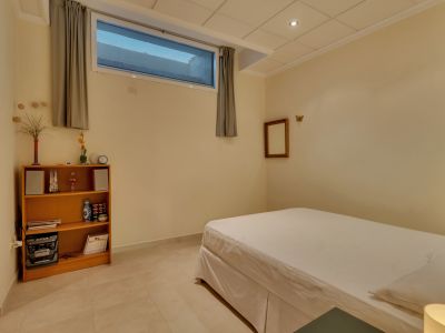 Habitación secundaria con cama individual, suelo de baldosas claras y paredes crema. Incluye una estantería de madera, un sistema de música y una ventana alta con cortinas grises.