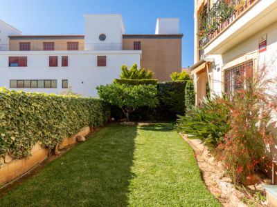 Jardín privado con césped bien cuidado, rodeado de exuberante vegetación y un pequeño árbol frutal. Se aprecian edificios modernos al fondo y una fachada con toldo.