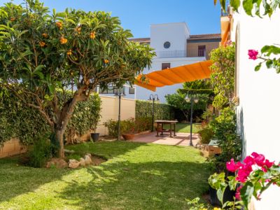 Jardín privado con césped, un níspero frutal y setos verdes. Patio pavimentado con mesa de madera bajo toldo naranja retráctil, complementado con iluminación exterior y bougainvillea rosa.