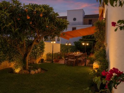 Jardín privado iluminado al anochecer con níspero, mesa de comedor exterior bajo toldo naranja y exuberante vegetación.
