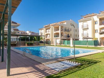 Piscina comunitaria de agua salada con azulejos azules, césped verde y edificios residenciales en el fondo. Pérgola de madera y ducha exterior.
