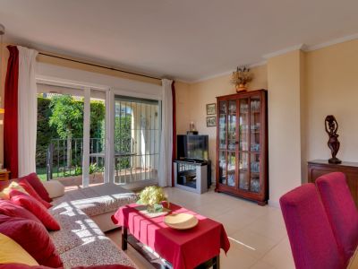 Sala de estar luminosa con sofá en L, cojines rojos y amarillos. Acceso directo a terraza y jardín privado a través de puertas correderas, con mueble vitrina de madera y lámpara colgante.