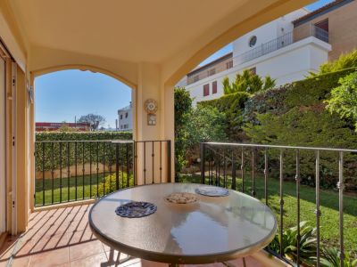 Terraza cubierta con mesa de cristal y suelo de terracota, con vistas directas a un jardín privado con césped y setos, y edificios distantes bajo luz natural.