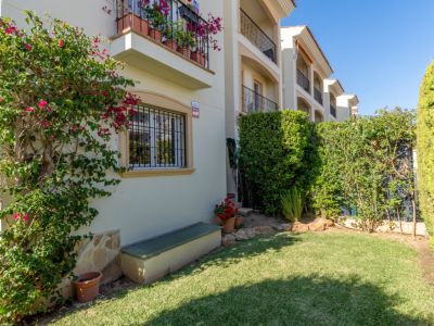 Vista del jardín privado con césped verde, buganvilla en flor en la fachada y setos. Incluye una ventana con rejas y un banco exterior.