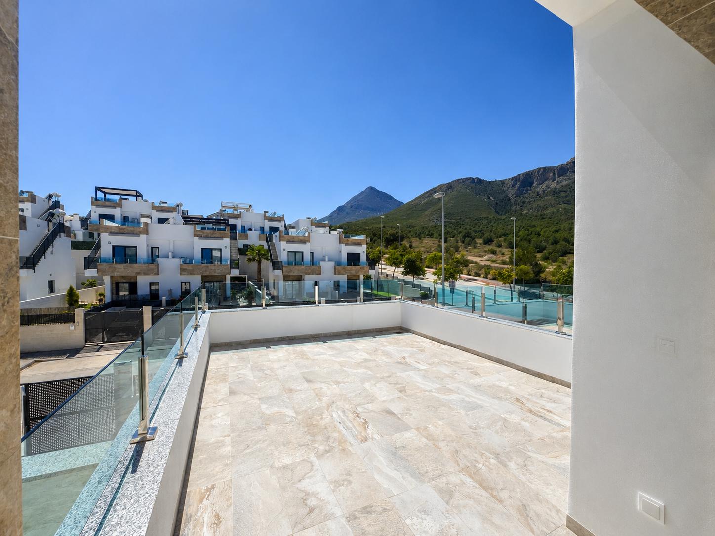 Amplia terraza exterior con suelo de gres claro, barandilla de cristal y vistas a edificios residenciales modernos y montañas cubiertas de vegetación bajo un cielo azul claro.