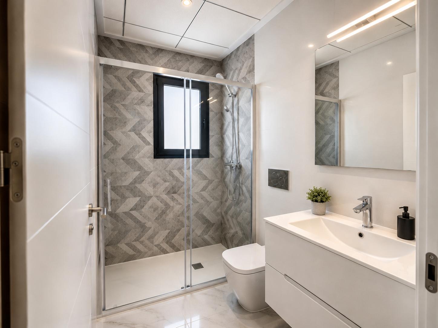Baño moderno con ducha walk-in, azulejos geométricos en tonos grises y ventana. Mueble de lavabo blanco, espejo iluminado y suelo de mármol.