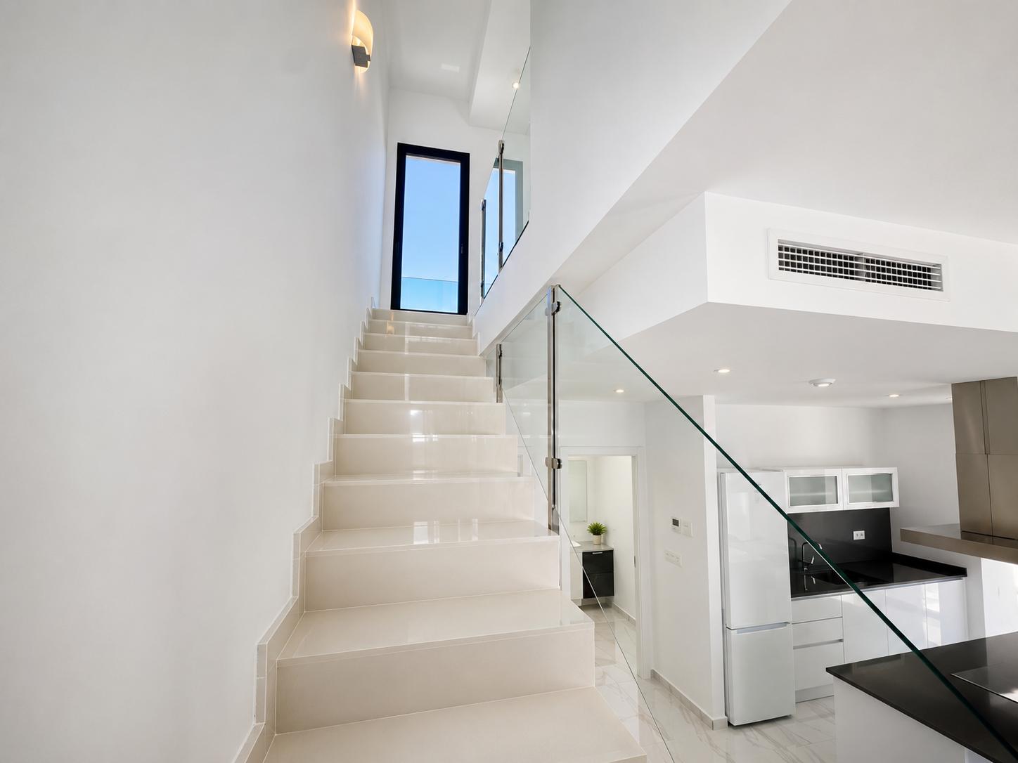 Escalera de diseño moderno con peldaños claros y barandilla de cristal, que conduce a una puerta exterior. Se visualiza parte de la cocina con encimeras negras y un baño auxiliar.