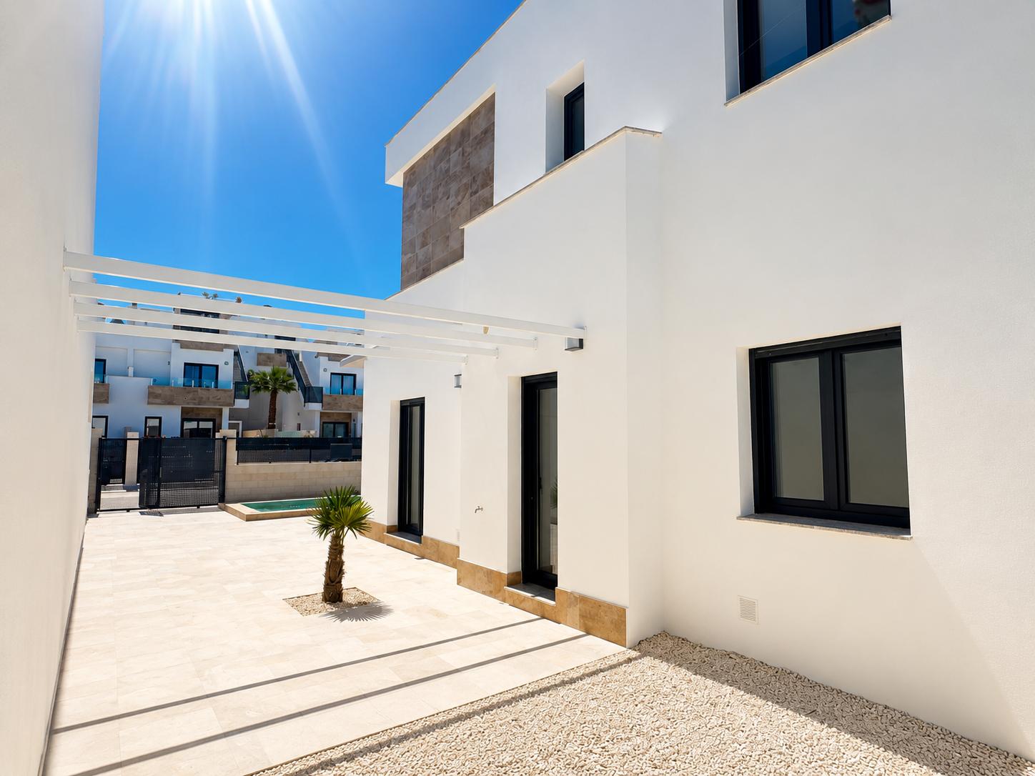 Exterior de villa moderna con fachada blanca y detalles de piedra. Terraza pavimentada con una pequeña palmera, pérgola blanca y ventanas de marco oscuro.