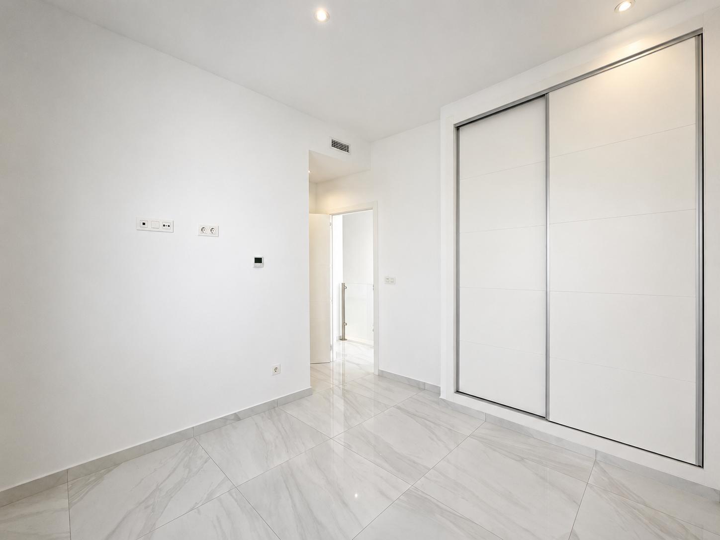 Habitación moderna con amplios armarios empotrados blancos de puertas correderas, suelo de gres porcelánico efecto mármol y focos empotrados en el techo. Un diseño limpio y funcional.