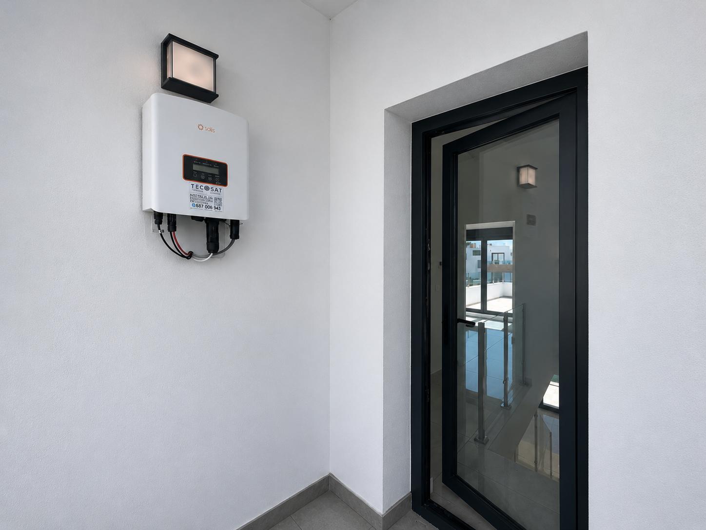Inversor solar Solis montado en pared blanca, con luminaria exterior. Puerta de vidrio con marco negro semiabierta que muestra una escalera interior con barandilla.