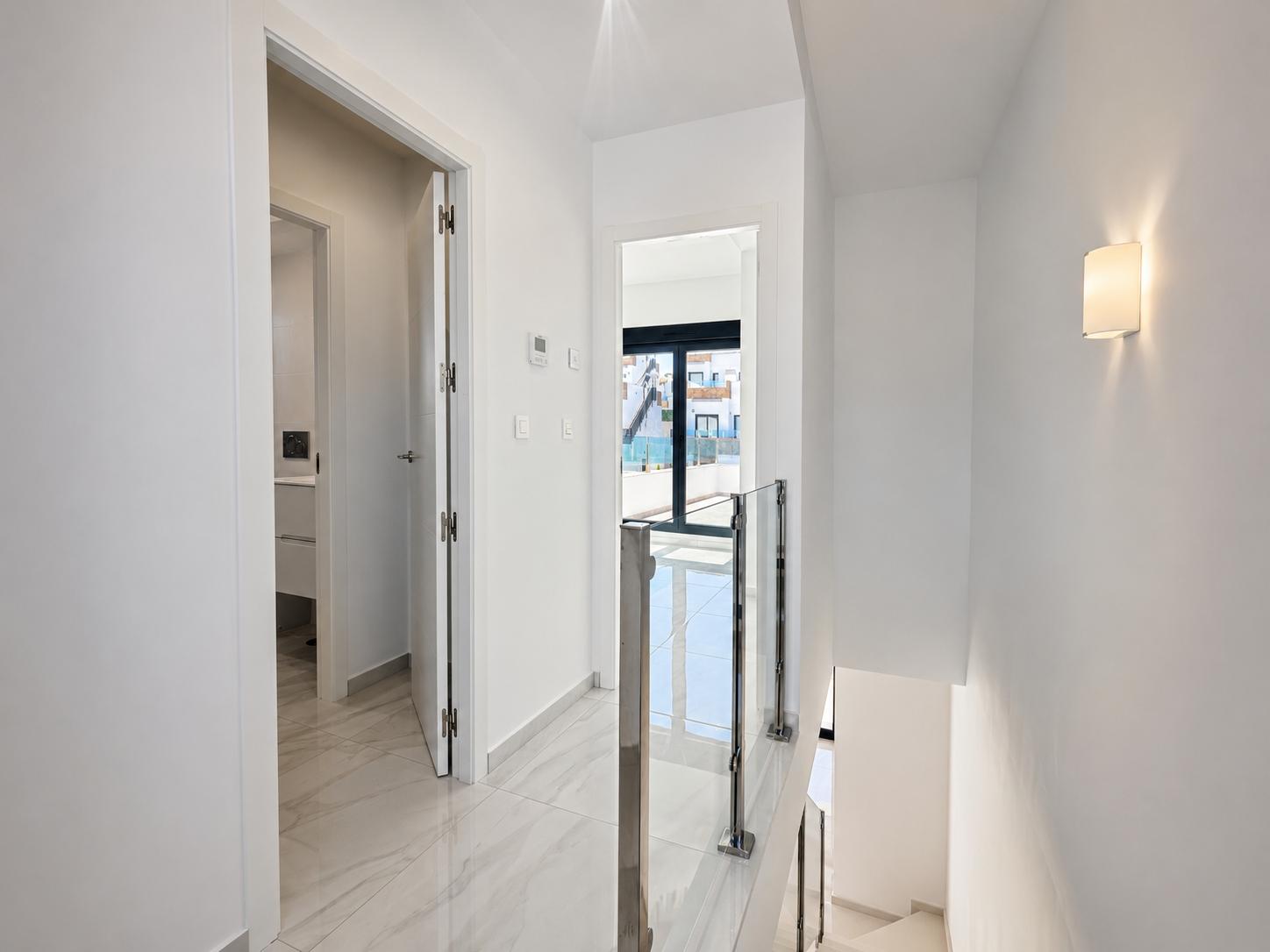Luminoso pasillo superior con suelos de gres porcelánico efecto mármol y barandilla moderna de cristal y acero. Una puerta conduce a un baño, otra a un balcón exterior con luz natural.