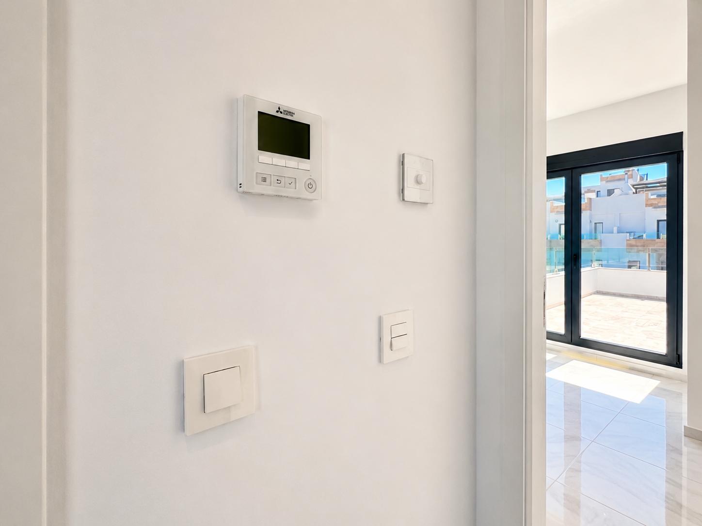 Panel de control de aire acondicionado Mitsubishi y modernos interruptores en pared blanca, junto a grandes puertas correderas que dan a una terraza alicatada con barandilla de cristal.