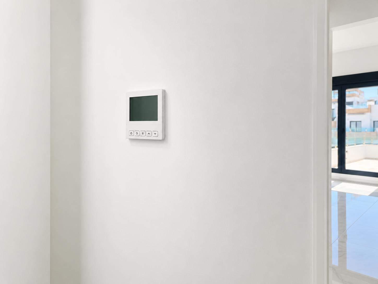 Panel de control digital moderno para la climatización en pared blanca, junto a un espacio interior luminoso con grandes ventanales y suelo reflectante.