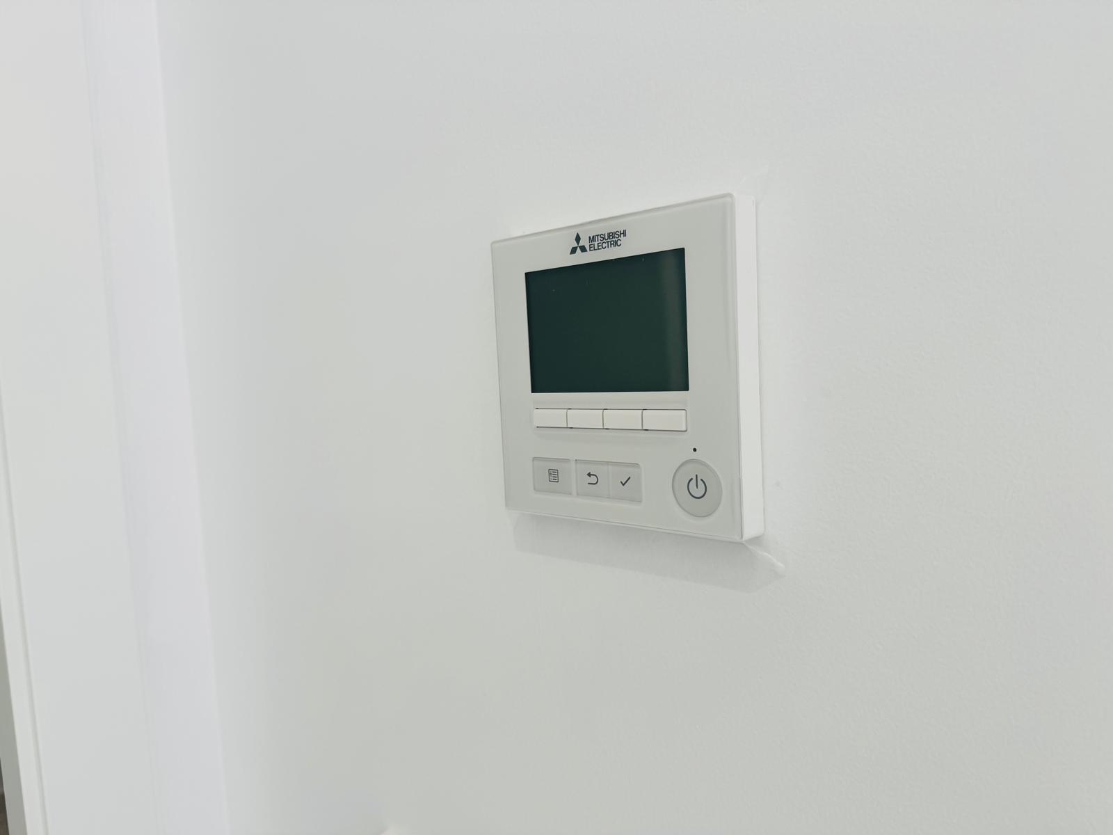Panel de control Mitsubishi Electric para aire acondicionado por conductos, con pantalla digital y botones intuitivos, integrado en la pared blanca.