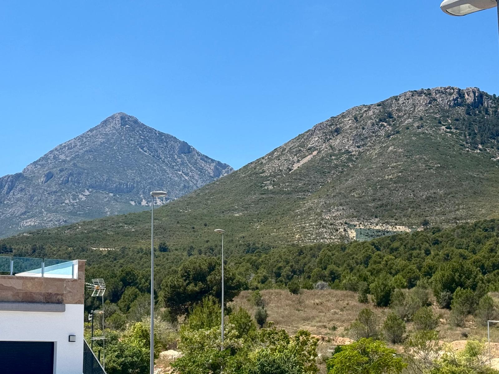 Vista despejada a dos montañas rocosas y abundante vegetación mediterránea, con una moderna terraza y barandilla de cristal en primer plano.