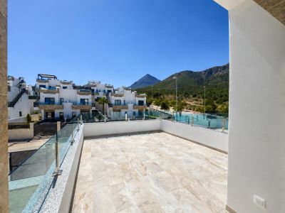 Amplia terraza exterior con suelo de gres claro, barandilla de cristal y vistas a edificios residenciales modernos y montañas cubiertas de vegetación bajo un cielo azul claro.