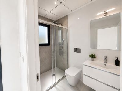 Baño moderno con lavabo flotante blanco, inodoro suspendido y ducha a ras de suelo con azulejos grises texturizados y ventana esmerilada. Suelo de gres porcelánico efecto mármol.