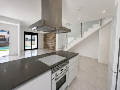 Cocina americana con isla de encimera oscura, placa integrada y horno. El espacio abierto conecta con el salón y la piscina, destacando una escalera de vidrio y pared de piedra.