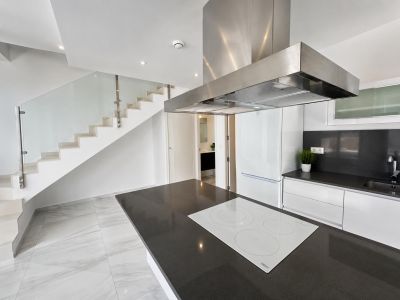 Cocina americana con isla de encimera oscura y placa de inducción blanca, junto a una escalera moderna con barandilla de cristal y peldaños blancos.