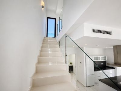 Escalera de diseño moderno con peldaños claros y barandilla de cristal, que conduce a una puerta exterior. Se visualiza parte de la cocina con encimeras negras y un baño auxiliar.