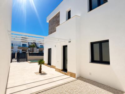 Exterior de villa moderna con fachada blanca y detalles de piedra. Terraza pavimentada con una pequeña palmera, pérgola blanca y ventanas de marco oscuro.