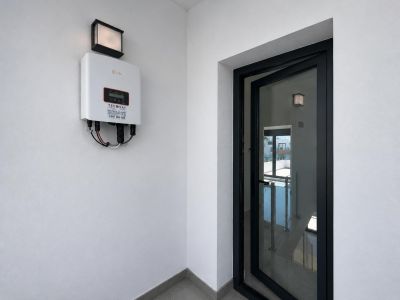 Inversor solar Solis montado en pared blanca, con luminaria exterior. Puerta de vidrio con marco negro semiabierta que muestra una escalera interior con barandilla.