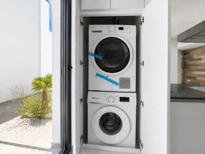Lavadora y secadora integradas en armario blanco, con acceso directo a un patio exterior ajardinado. Al fondo, se aprecia una moderna cocina con pared de madera y encimera oscura.