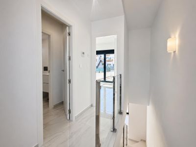 Luminoso pasillo superior con suelos de gres porcelánico efecto mármol y barandilla moderna de cristal y acero. Una puerta conduce a un baño, otra a un balcón exterior con luz natural.
