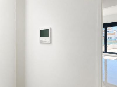 Panel de control digital moderno para la climatización en pared blanca, junto a un espacio interior luminoso con grandes ventanales y suelo reflectante.