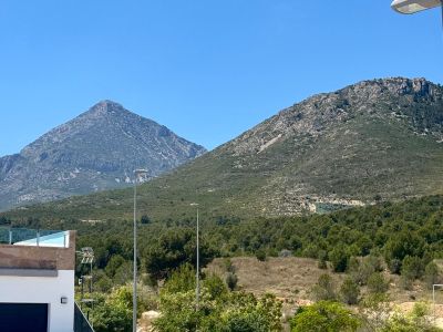 Vista despejada a dos montañas rocosas y abundante vegetación mediterránea, con una moderna terraza y barandilla de cristal en primer plano.