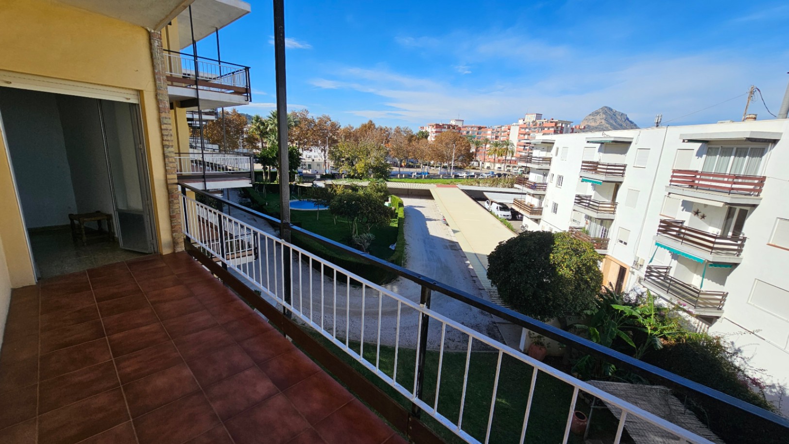 Amplio balcón con suelo de baldosas de terracota, ofreciendo vistas al jardín comunitario con piscina, edificios residenciales y una montaña lejana bajo un cielo despejado.