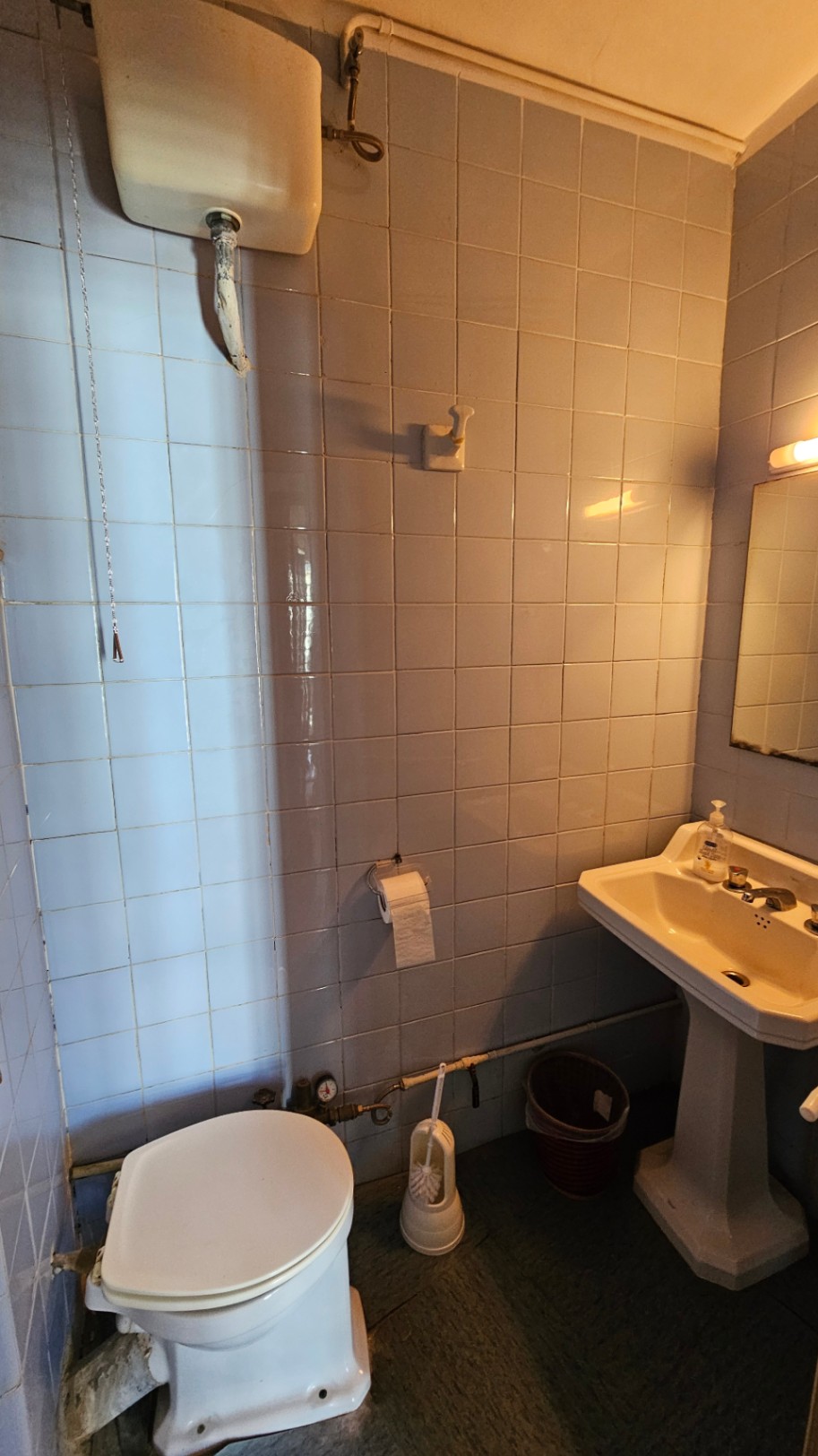 Baño compacto con azulejos cuadrados azul claro, cisterna alta con cadena, lavabo de pedestal y tuberías vistas. Incluye espejo y aplique de pared.