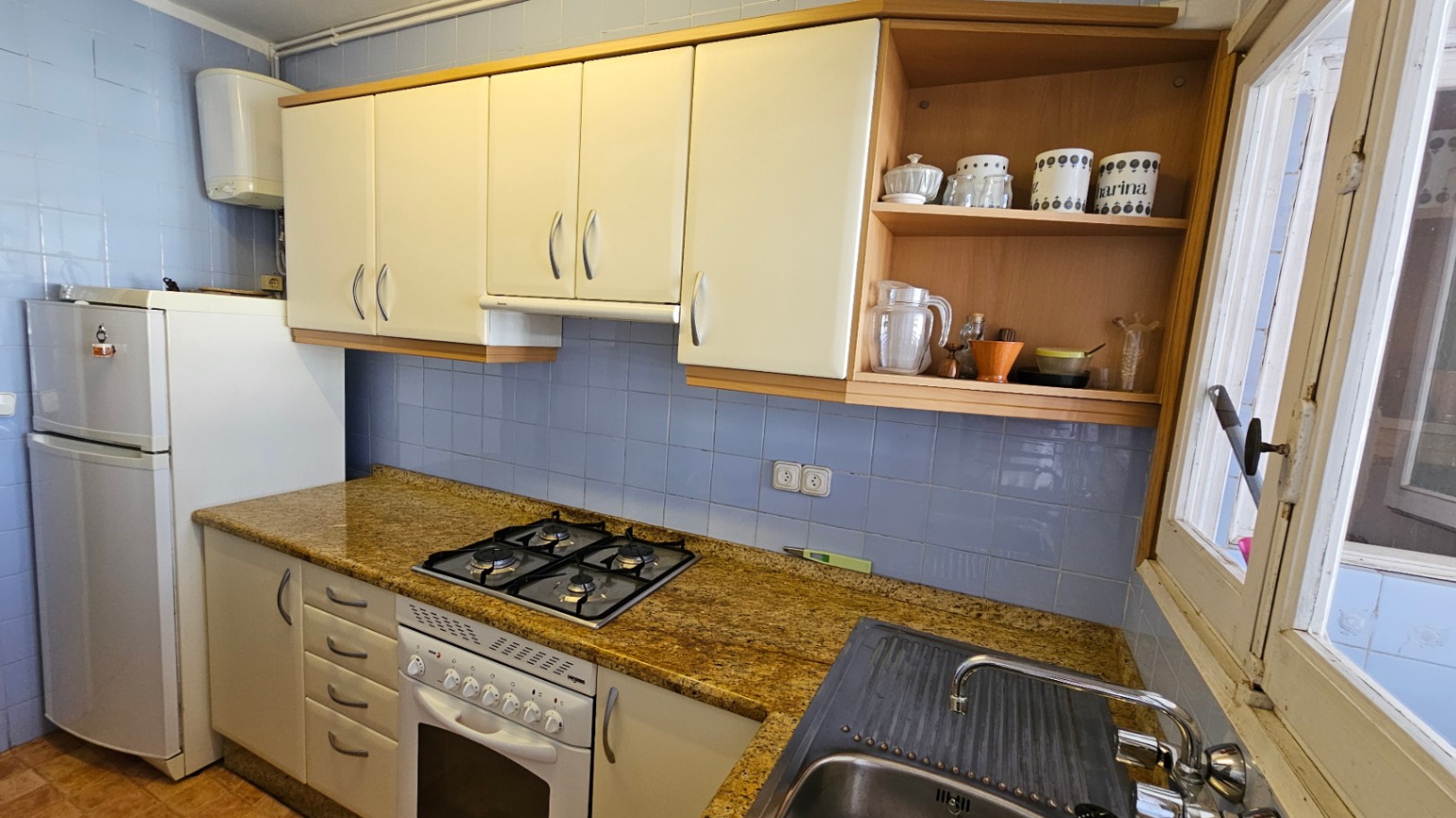 Cocina funcional con azulejos azules, encimera de granito moteado y mobiliario en tonos crema. Incluye placa de gas, horno y fregadero de acero, con luz natural de la ventana.