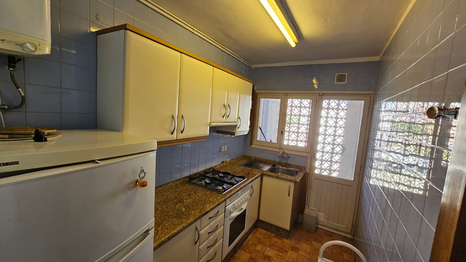 Cocina funcional con azulejos azules, encimera de granito y armarios blancos. Dispone de horno, fogones de gas y luz natural a través de una ventana con celosía.
