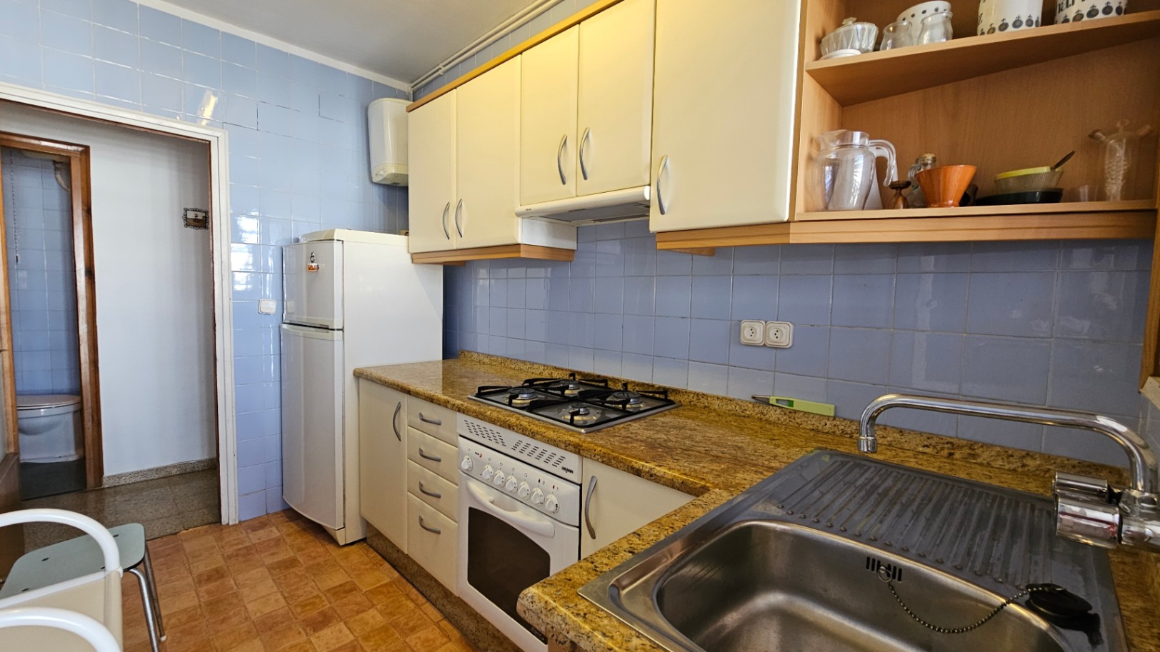 Cocina funcional con azulejos azules, encimera efecto granito y armarios blancos. Equipada con placa de gas, horno, fregadero de acero inoxidable y nevera.