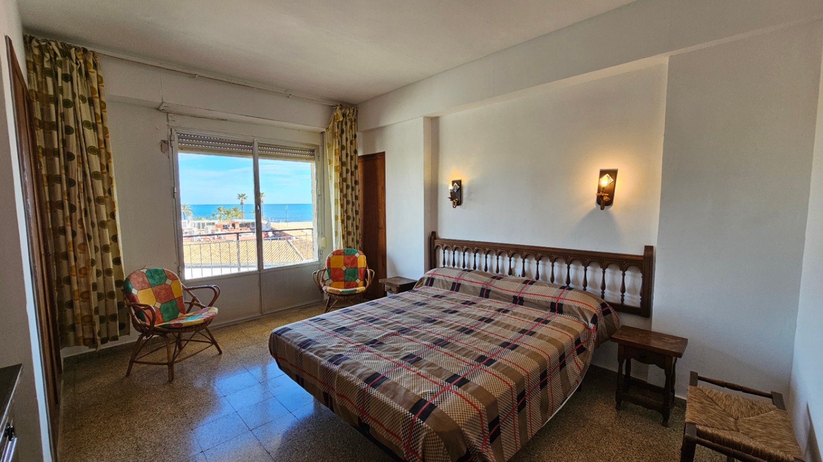 Dormitorio con cama de matrimonio y cabecero de madera tallada, ofreciendo vistas directas al mar Mediterráneo desde su ventana con persiana enrollable y cortinas estampadas. Suelo de terrazo.
