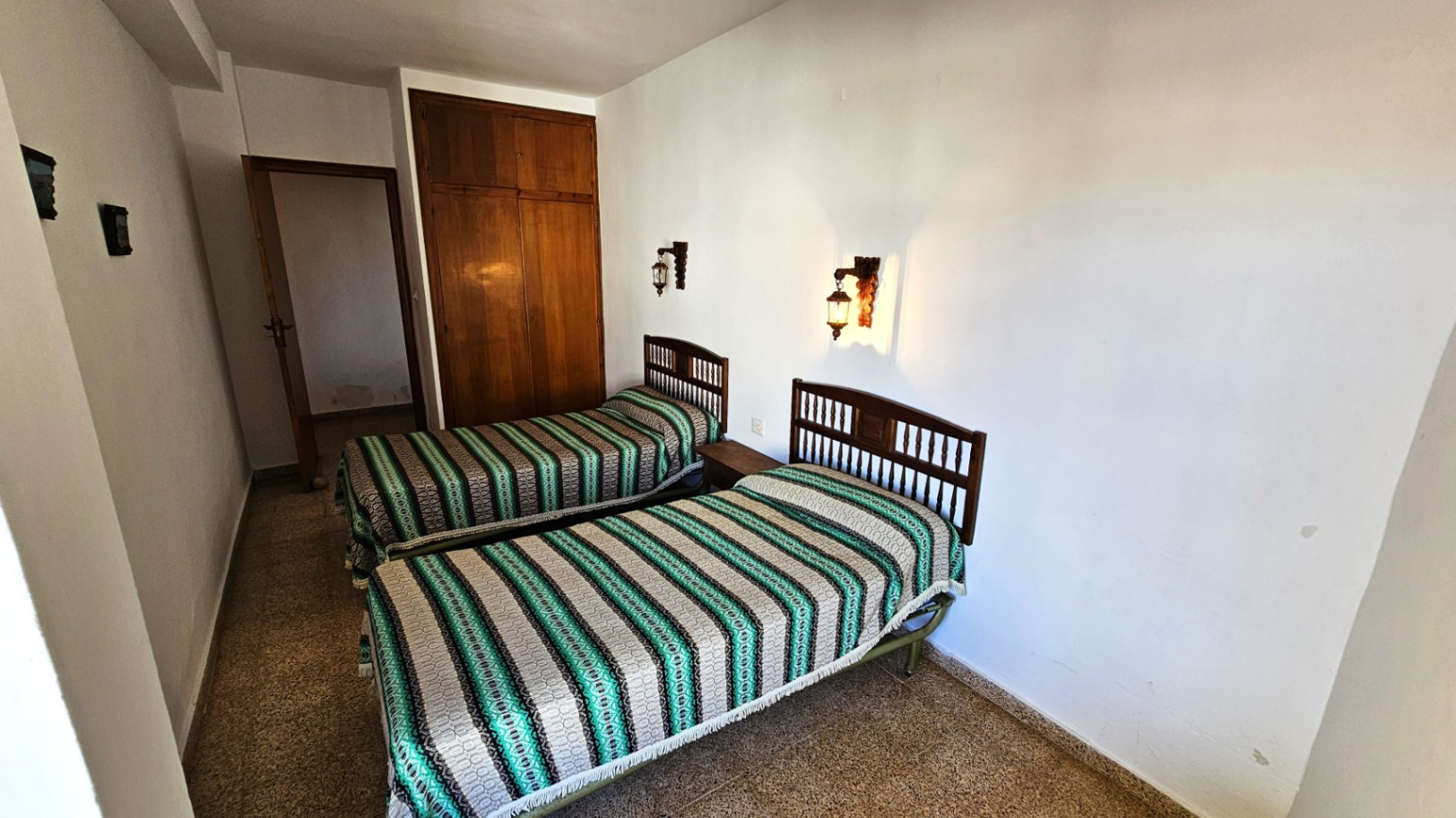 Dormitorio con dos camas individuales de cabecero de madera y colchas a rayas. Incluye armario empotrado de madera y apliques de pared, sobre un suelo de gres.