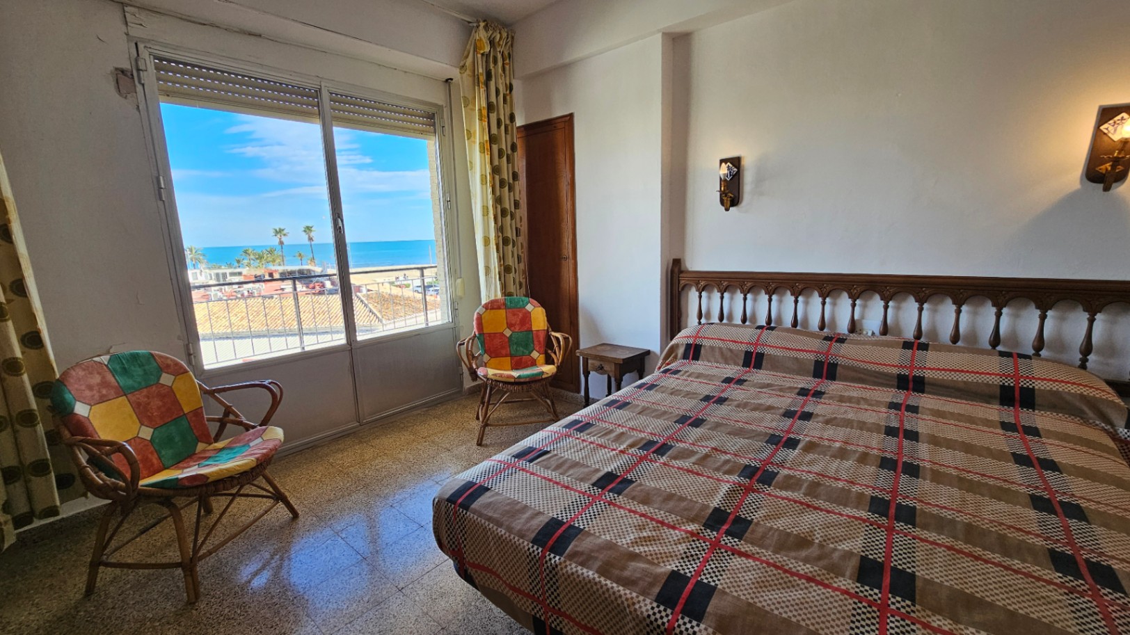 Dormitorio con vistas directas al mar desde un gran ventanal, cama con cabecero de madera, colcha a cuadros y dos sillas de ratán. Luz natural sobre el suelo de gres.