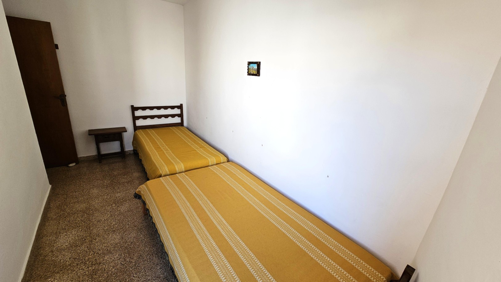 Dormitorio sencillo con dos camas individuales y colchas amarillas a rayas. Incluye un cabecero de madera oscura, una mesita de noche y suelo de terrazo.