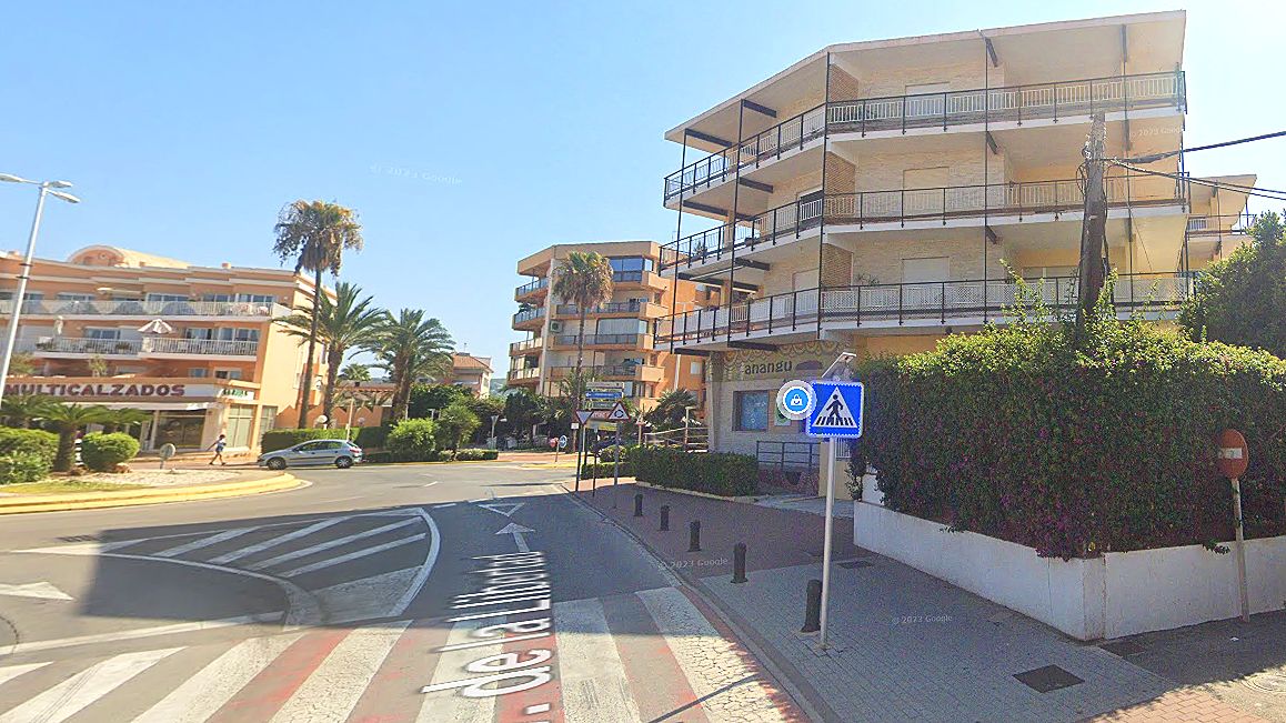 Edificio residencial en esquina con fachada clara y múltiples balcones, situado junto a un cruce de calles con paso de peatones y vegetación.
