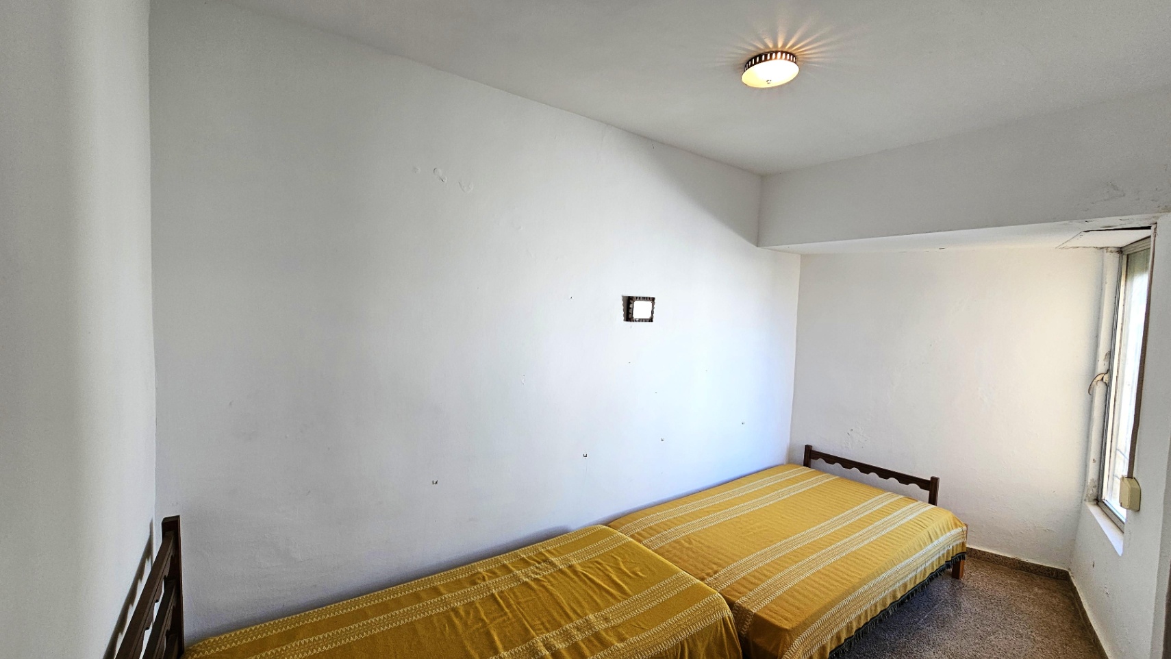 Habitación con dos camas individuales y cabeceros de madera. Las paredes blancas muestran signos de uso, con una ventana lateral que aporta luz natural y un suelo de terrazo.