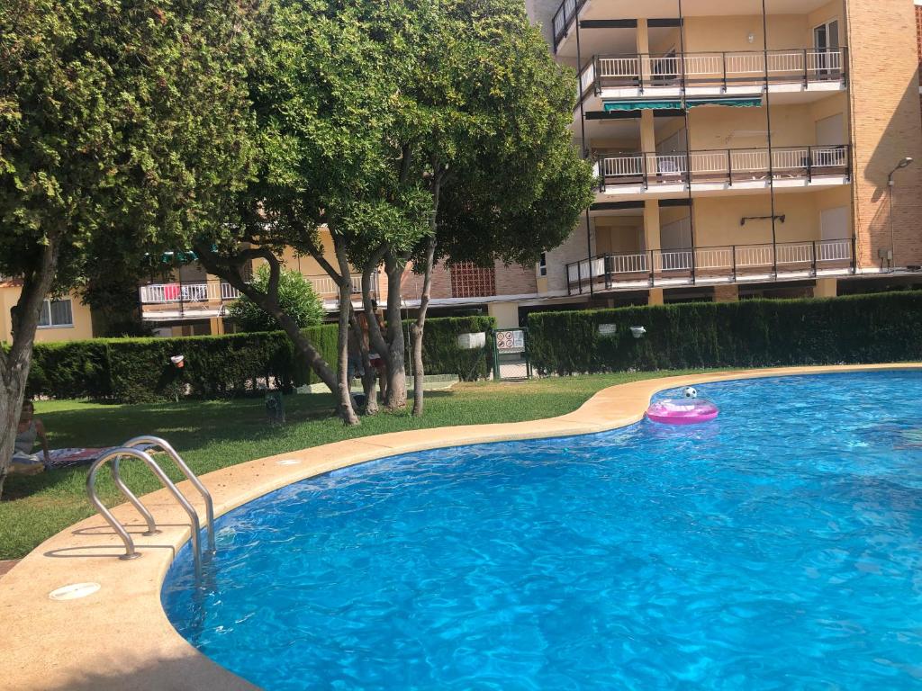 Piscina comunitaria con borde curvo de hormigón y escaleras metálicas, rodeada de césped y árboles maduros. Edificio residencial con balcones visible al fondo.