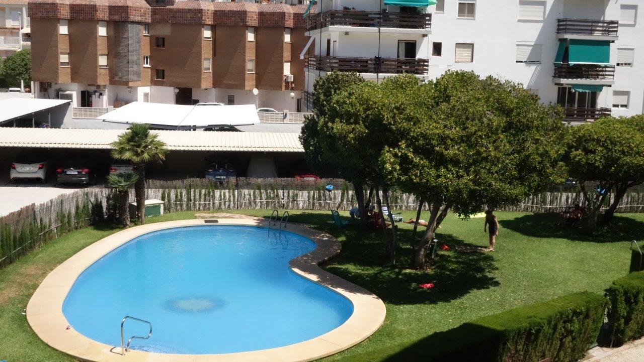 Piscina comunitaria de forma ovalada con bordillo claro, rodeada de césped verde y árboles que ofrecen sombra natural. Al fondo, aparcamiento cubierto y edificios residenciales.