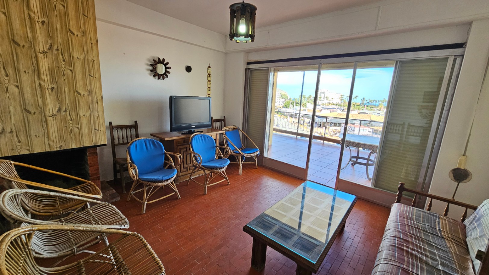 Sala de estar con suelo de terracota, chimenea con revestimiento de madera vertical y mobiliario de mimbre. Amplias puertas correderas dan a una terraza exterior con vistas urbanas.