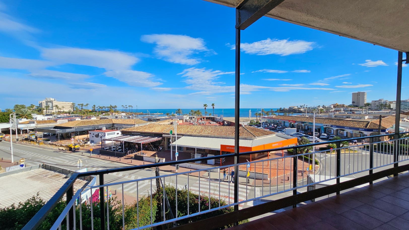 Vista desde la terraza sobre una calle animada con comercios y tejados de teja, extendiéndose hacia la Playa del Arenal y el azul claro del Mediterráneo bajo un cielo brillante.