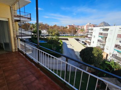 Amplio balcón con suelo de baldosas de terracota, ofreciendo vistas al jardín comunitario con piscina, edificios residenciales y una montaña lejana bajo un cielo despejado.
