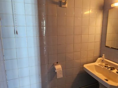 Baño compacto con azulejos cuadrados azul claro, cisterna alta con cadena, lavabo de pedestal y tuberías vistas. Incluye espejo y aplique de pared.