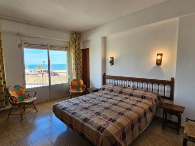 Dormitorio con cama de matrimonio y cabecero de madera tallada, ofreciendo vistas directas al mar Mediterráneo desde su ventana con persiana enrollable y cortinas estampadas. Suelo de terrazo.