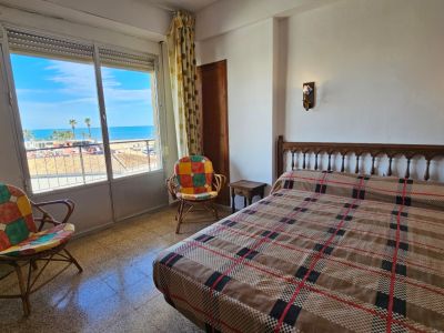 Dormitorio con vistas directas al mar desde un gran ventanal, cama con cabecero de madera, colcha a cuadros y dos sillas de ratán. Luz natural sobre el suelo de gres.