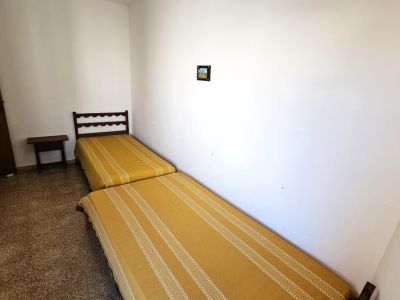 Dormitorio sencillo con dos camas individuales y colchas amarillas a rayas. Incluye un cabecero de madera oscura, una mesita de noche y suelo de terrazo.